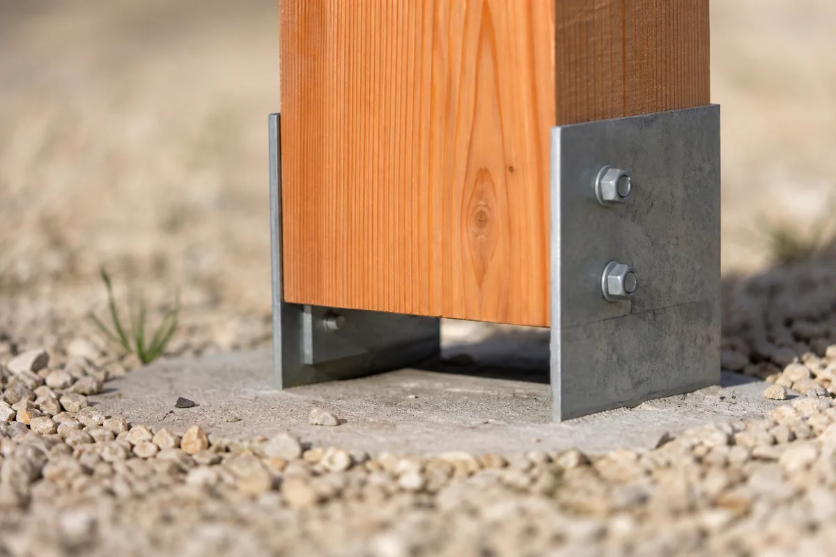 Détail d’un pied de poteau bois monté sur sabot métallique galvanisé déporté au-dessus d’un plot béton, sol gravillonné